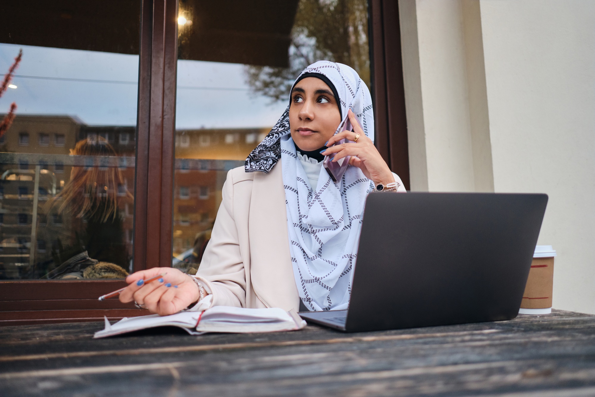 Young Arabic student in hijab confidently talking on smartphone working on laptop on city street